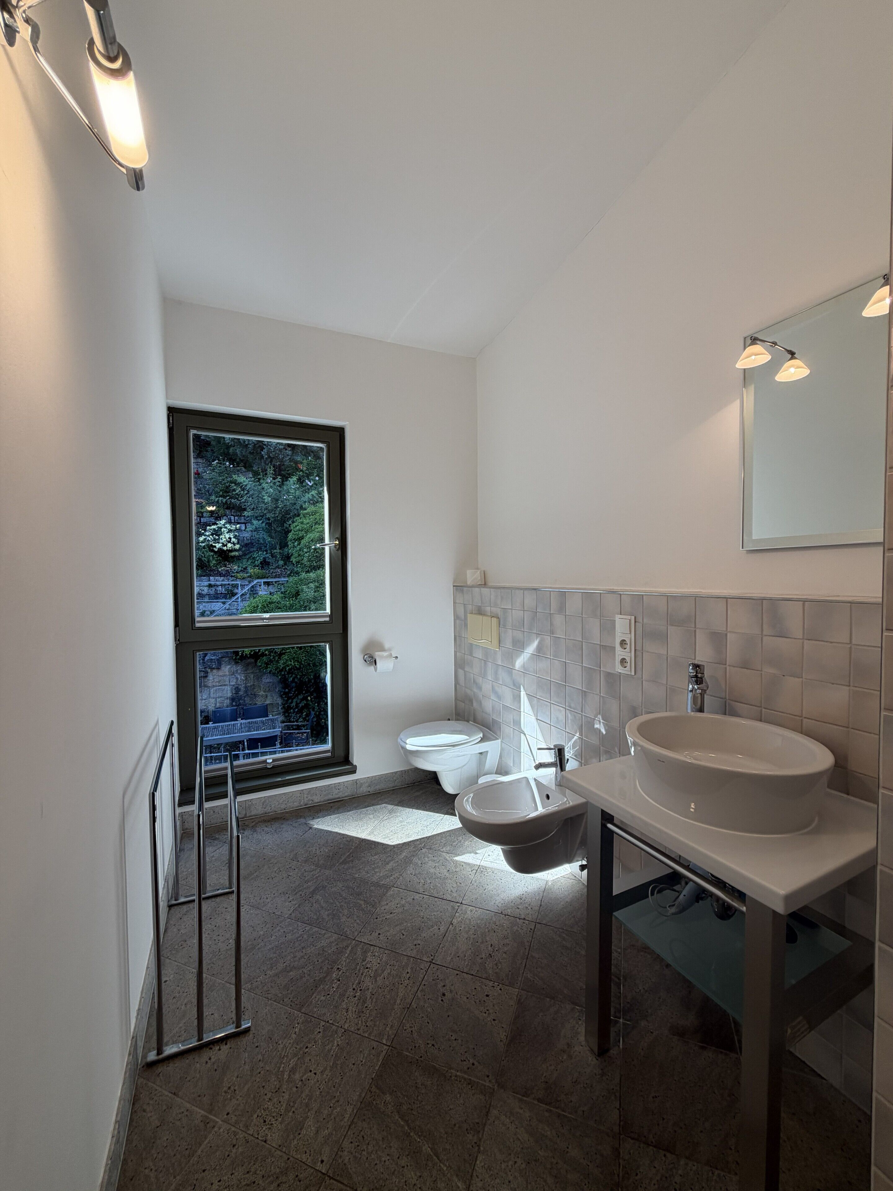 A modern bathroom with a large window, a toilet, a sink, and a mirror.