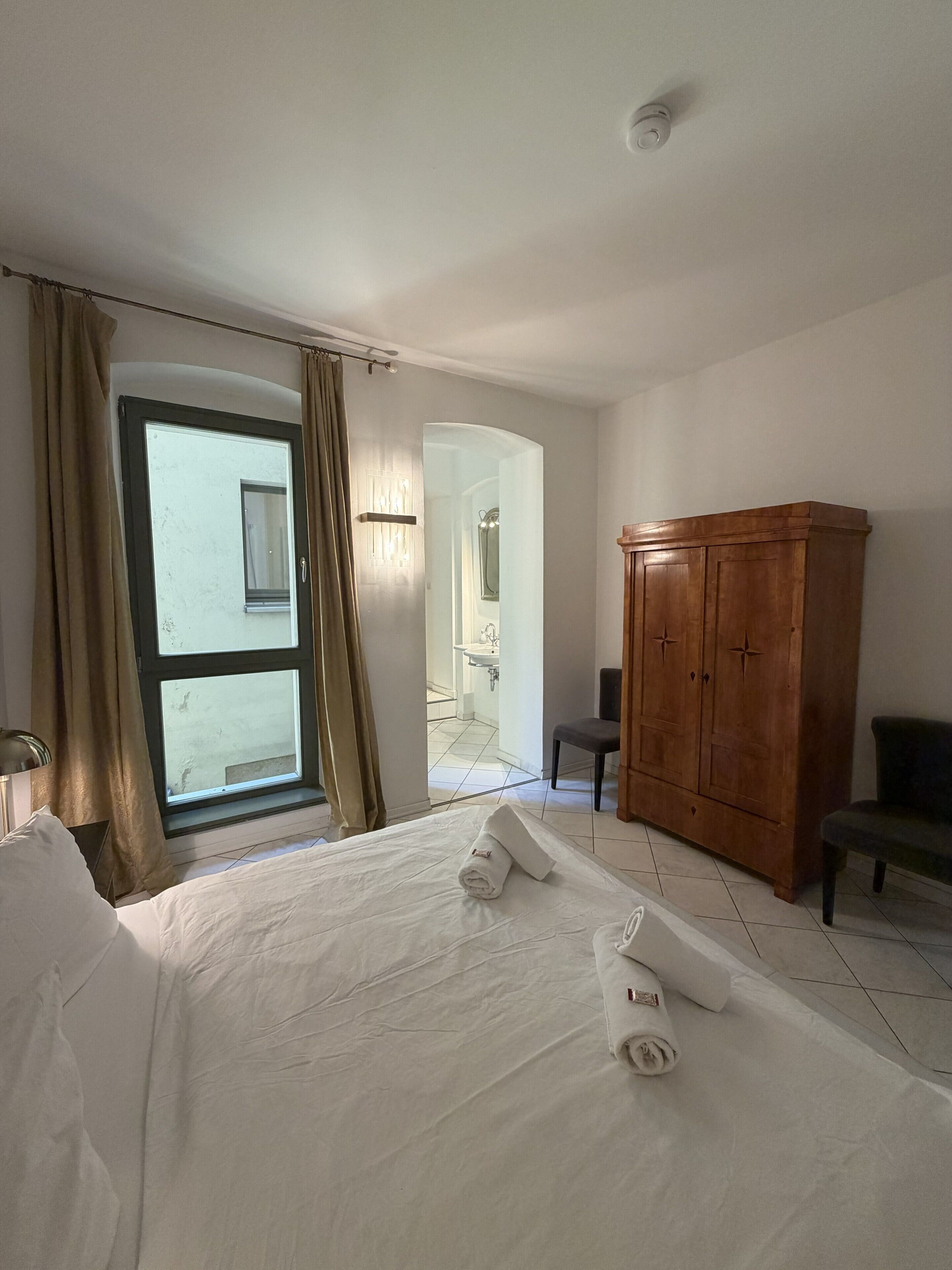 A hotel room with a bed, a wooden wardrobe, a chair, and a bathroom visible through an open door.