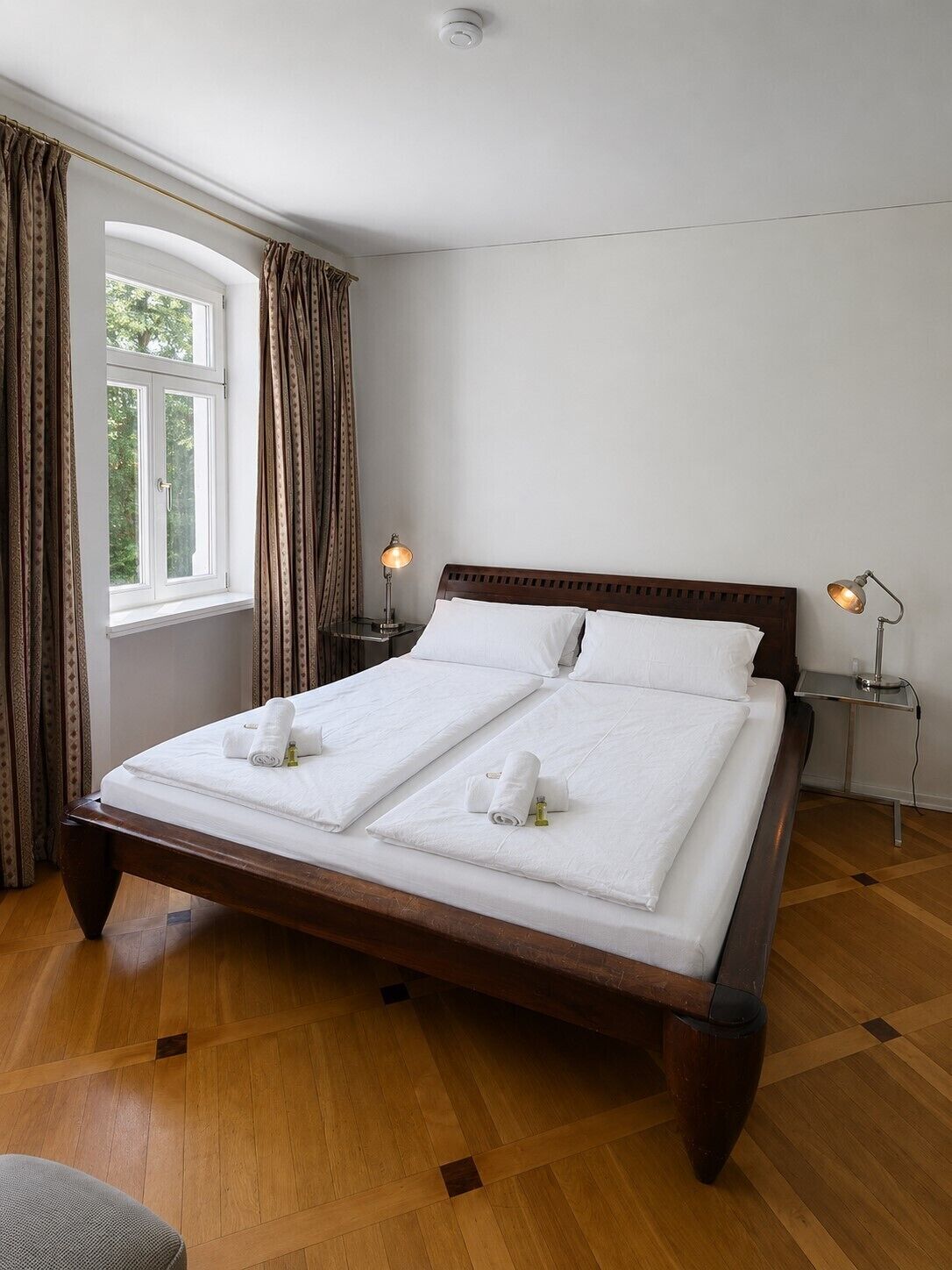 A double bed with white linens, two bedside tables with lamps, a window with curtains, and a wooden floor.