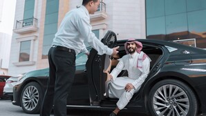 A man in traditional attire sitting in a car with a door open, and another person standing beside the car. - Julnar Hotel (Unaizah)