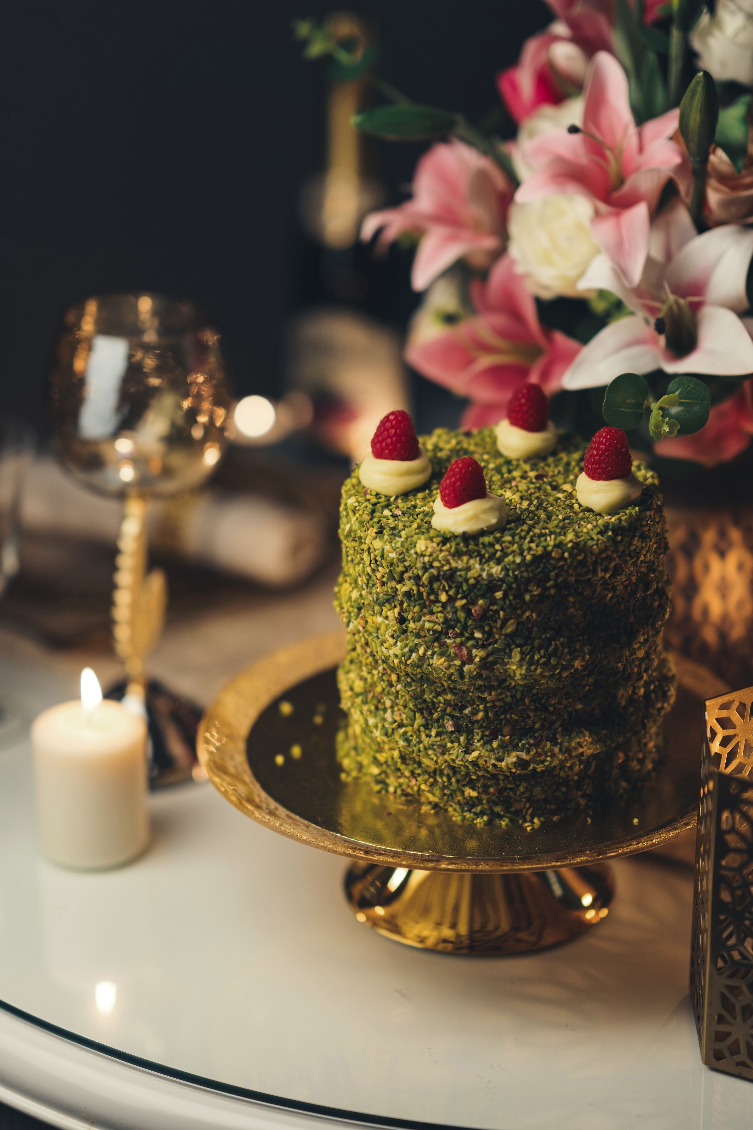 A cake on a golden stand with a candle and a glass of wine in the background.