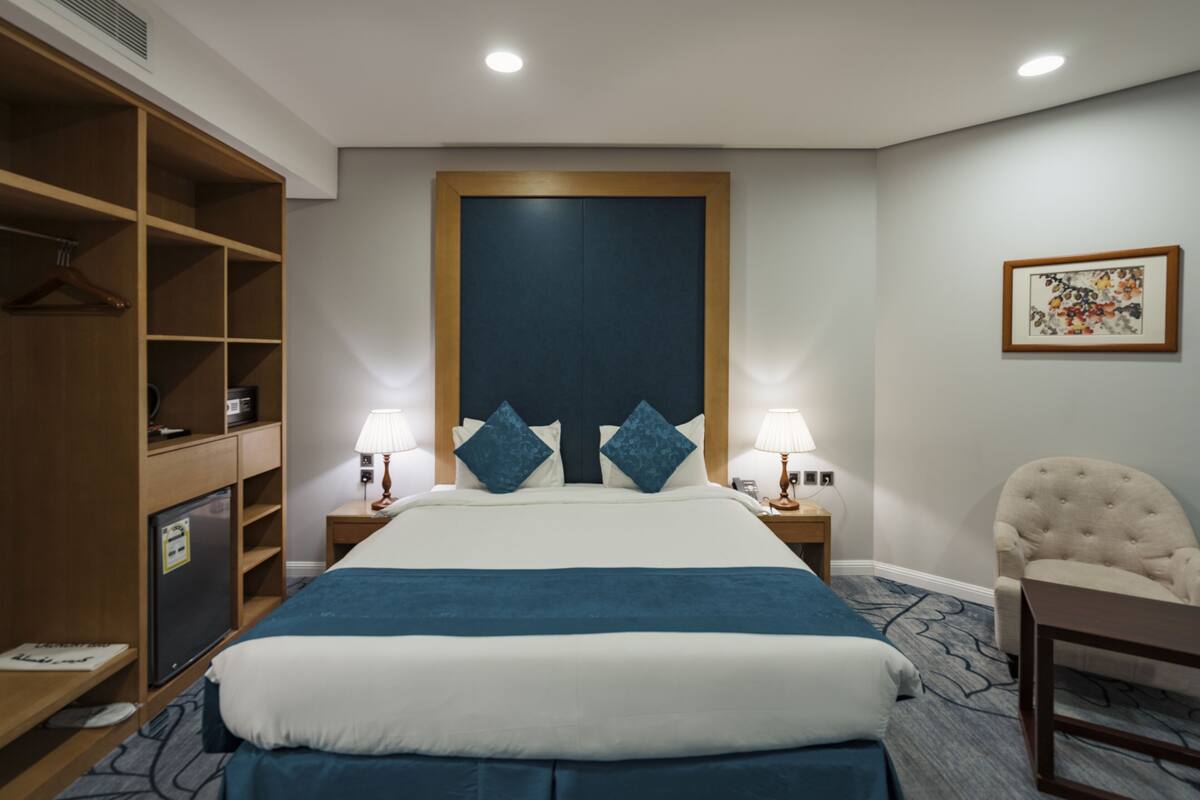 A hotel room with a large bed, two bedside tables with lamps, a wardrobe, a chair, and a framed picture on the wall.