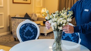 A person holding a vase of white flowers on a white table in a room with a blue and white patterned chair and a sofa in the background. - Julnar Hotel (Unaizah)