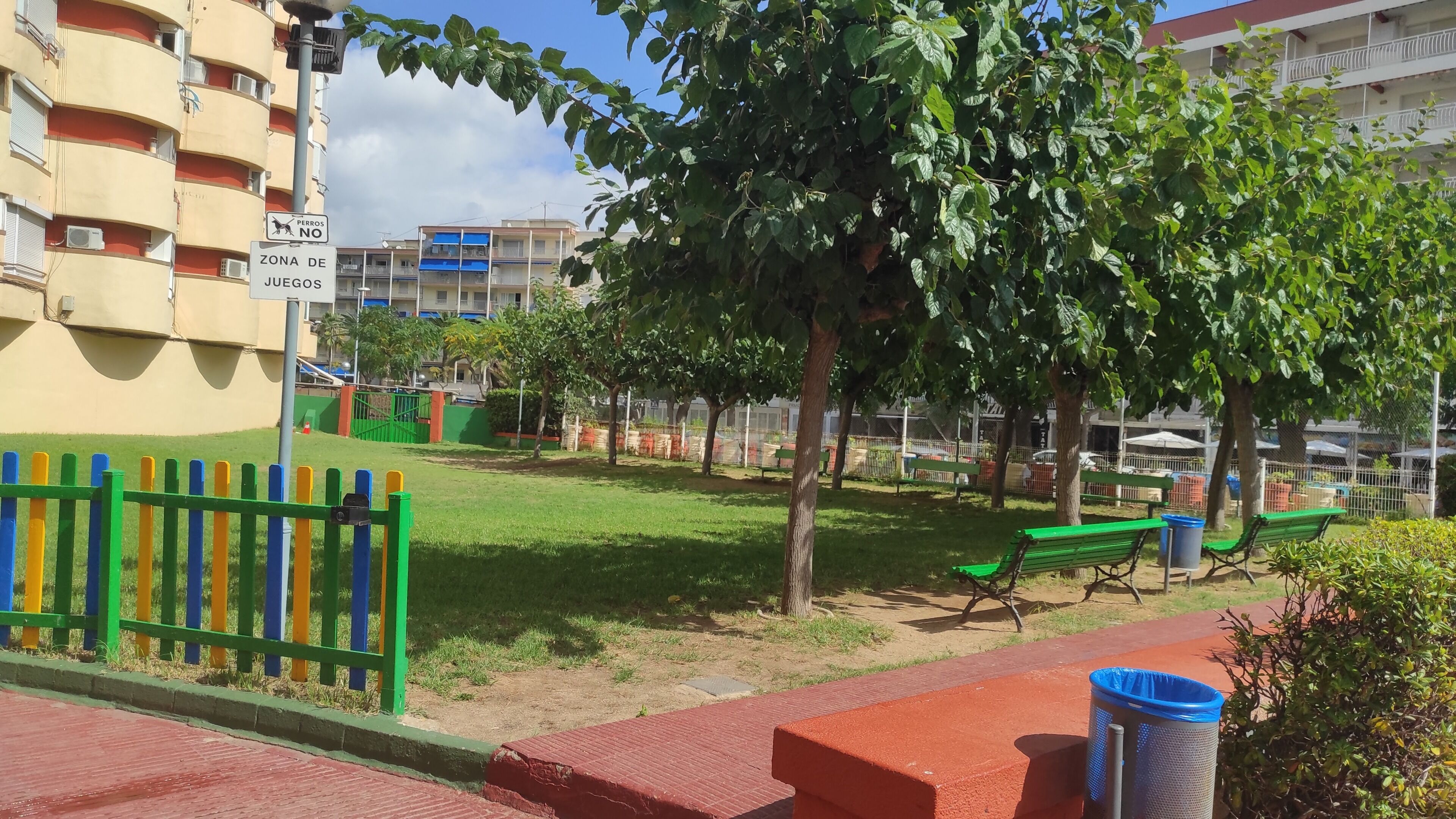 A park with benches, a trash bin, and a playground fence.