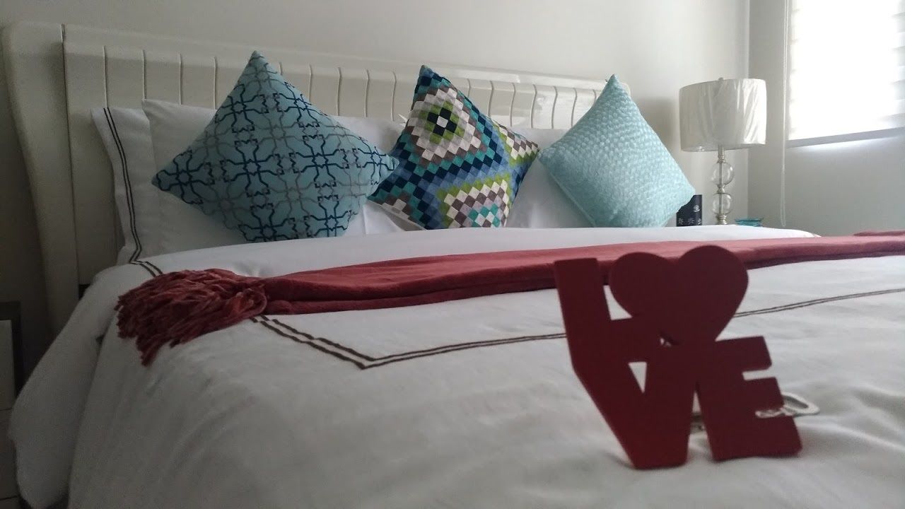 Een net opgemaakt bed met een witte deken, een rode plaid en een hartvormige decoratie. Er liggen drie kussens met verschillende patronen op de hoofdbord.