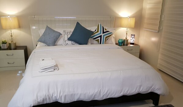 Una cama bien hecha con ropa de cama blanca, dos mesitas de noche con lámparas y una ventana con persianas.