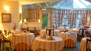 Ein Restaurant mit runden Tischen, bedeckt mit weißen Tischdecken und orangefarbenen Vorhängen, mit Glasdecke und Blick ins Freie.
