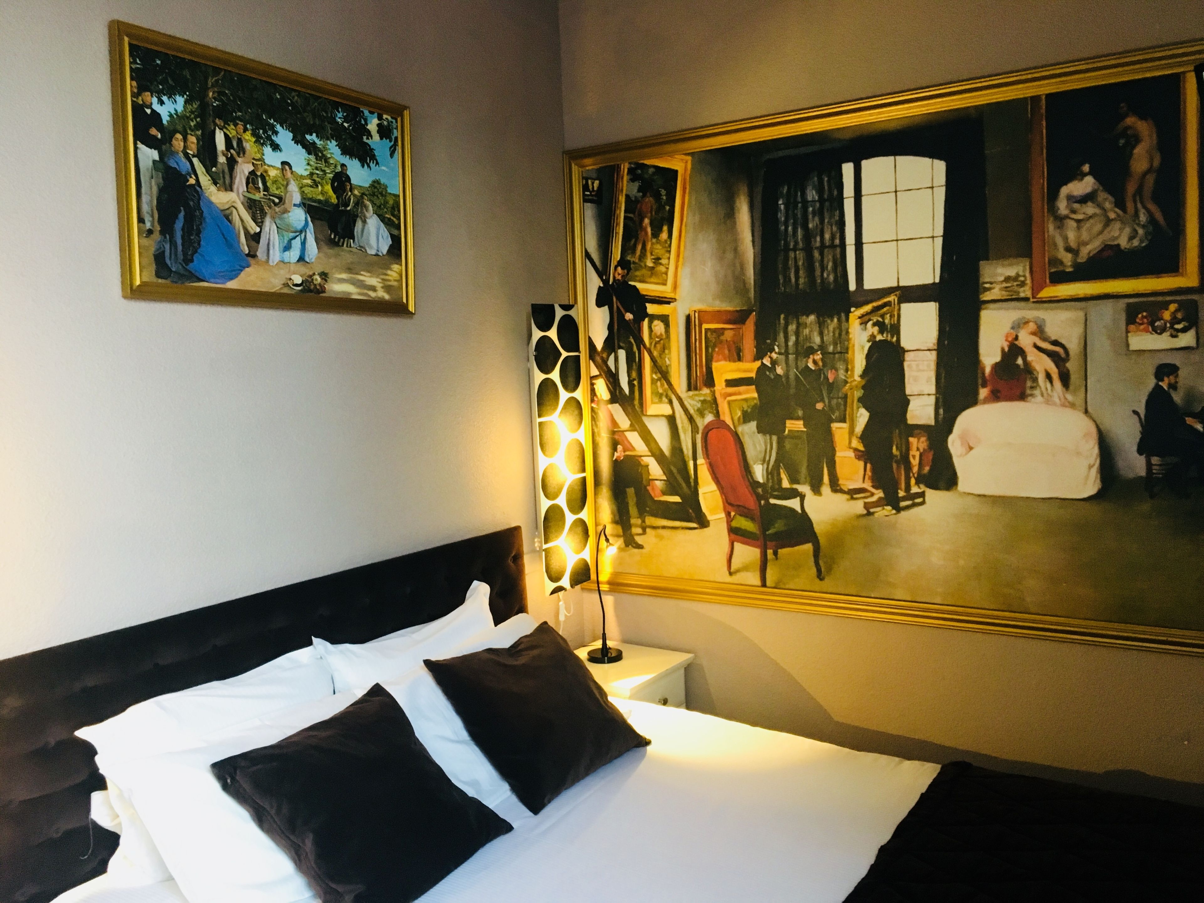 A hotel room with a large bed, a nightstand, a lamp, and two paintings on the wall.