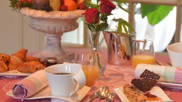 Une table de petit-déjeuner avec des croissants, une tasse de café, un saladier de fruits, un vase avec une rose et un verre de jus d’orange.