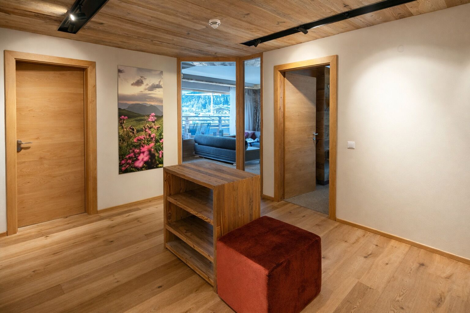 Un pasillo con suelo de madera, un reposapiés rojo, una estantería de madera y un cuadro en la pared.