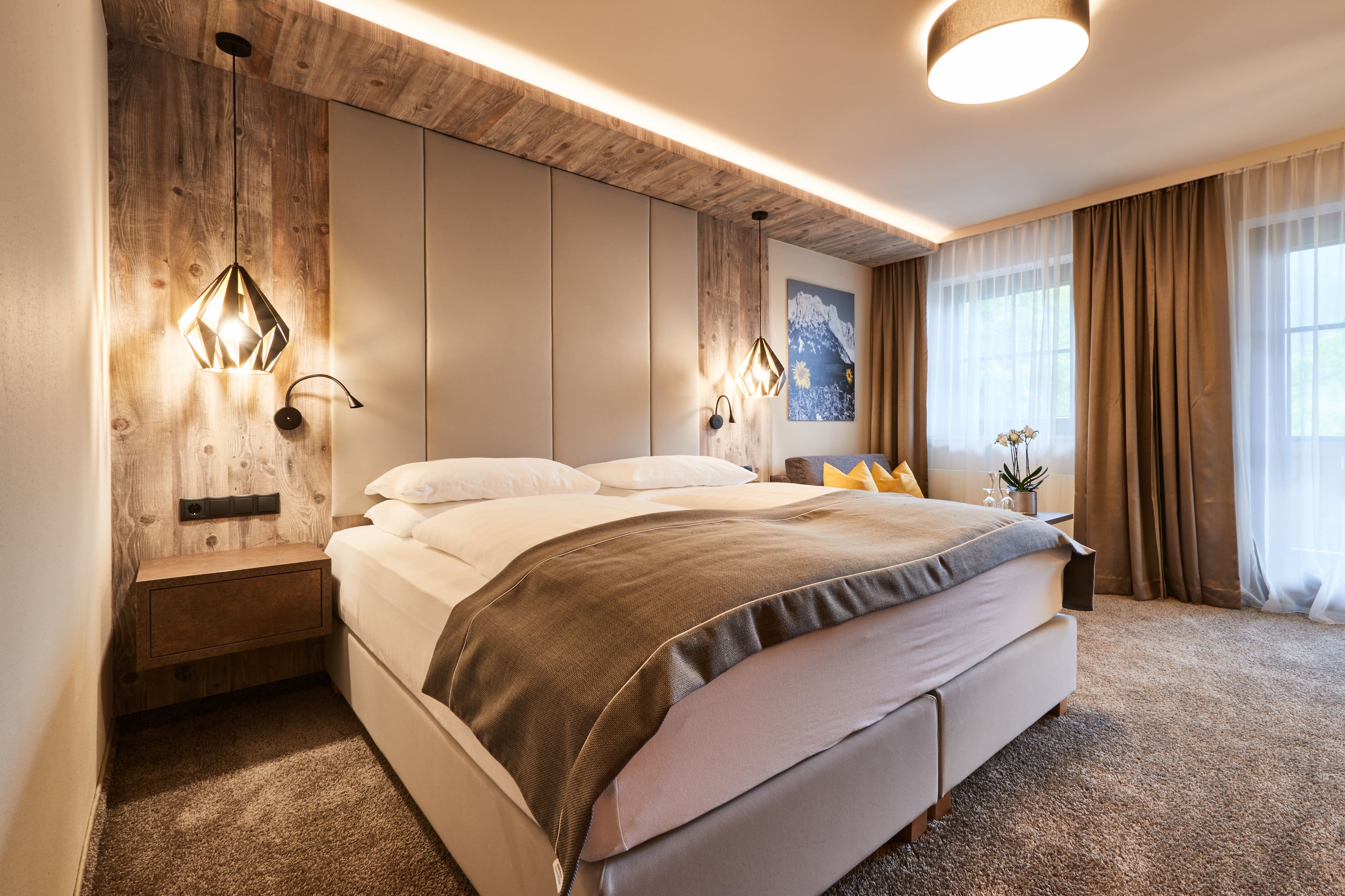 A modern bedroom with a wooden accent wall, a large bed, bedside lamps, and a view of the outdoors through sheer curtains.
