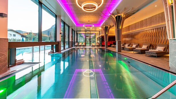 Piscina interior moderna com vista para montanhas, amplas janelas e área de lounge com assentos.