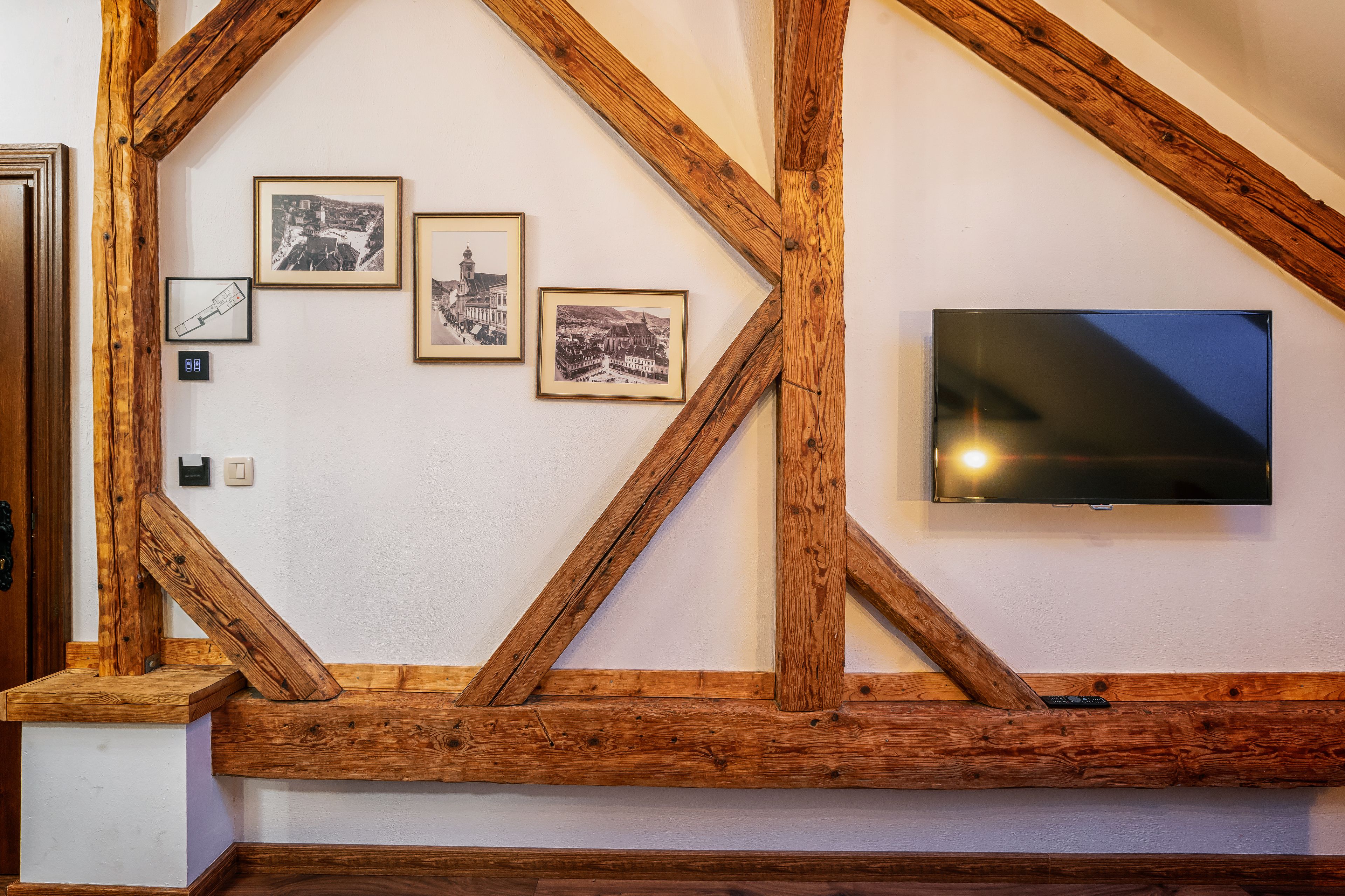 A room with exposed wooden beams, a mounted television, and three framed photographs on the wall.