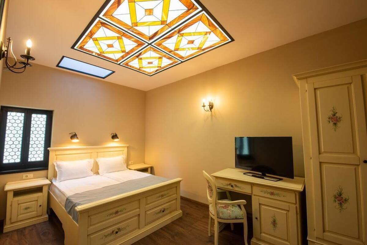 Quarto com cama, mesas de cabeceira, escrivaninha com cadeira, televisão e guarda-roupa.