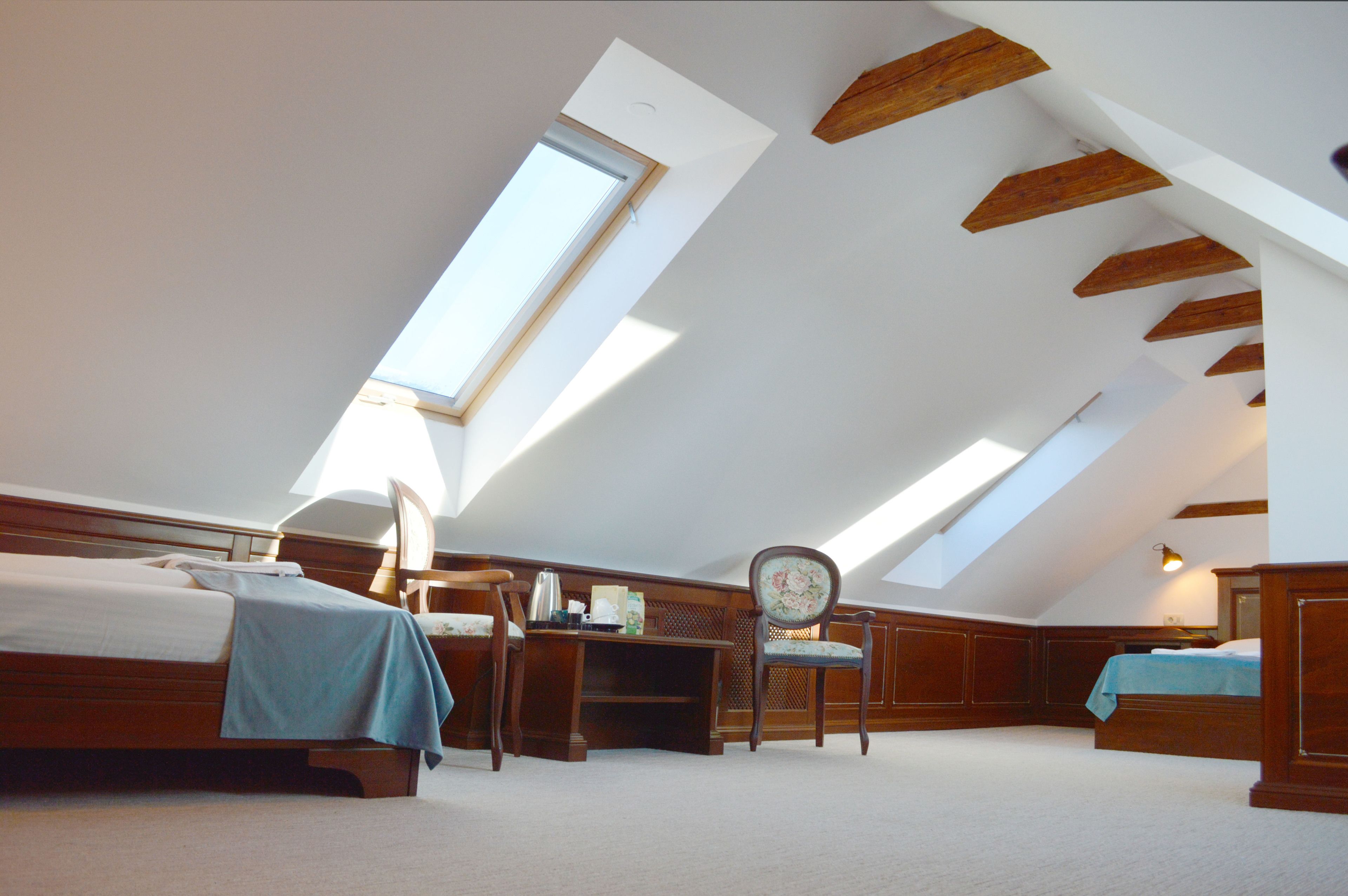 Ein Schlafzimmer mit schrägen Wänden, einem Oberlicht, Holzbalken, einem Bett, einem Schreibtisch, einem Stuhl und einem kleinen Tisch.
