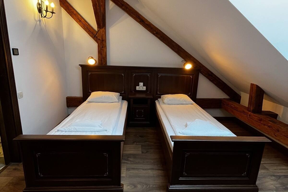 Quarto com duas camas de solteiro, vigas de madeira, teto inclinado e luminária fixada na parede.