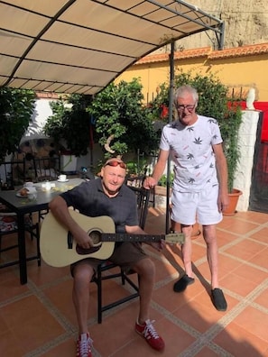 Two people sitting at an outdoor table with a guitar, under a shaded area with a tiled floor and potted plants. - Cuevaalegria (Guadix)