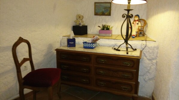 A room with a wooden chair, a desk with drawers, a lamp, and a small shelf with items.