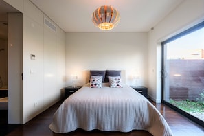 A modern bedroom with a large bed, two bedside tables, a unique pendant light, and a view through a glass door. - L'Orangerie (Vilamoura)