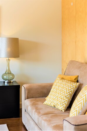 A beige sofa with two patterned pillows, a green lamp on a black side table, and a wooden wall. - L'Orangerie (Vilamoura)