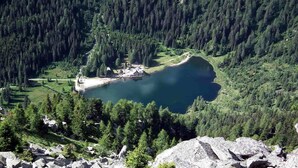 Paisaje montañoso con un lago rodeado de un bosque denso. Se ve una construcción pequeña cerca de la orilla.