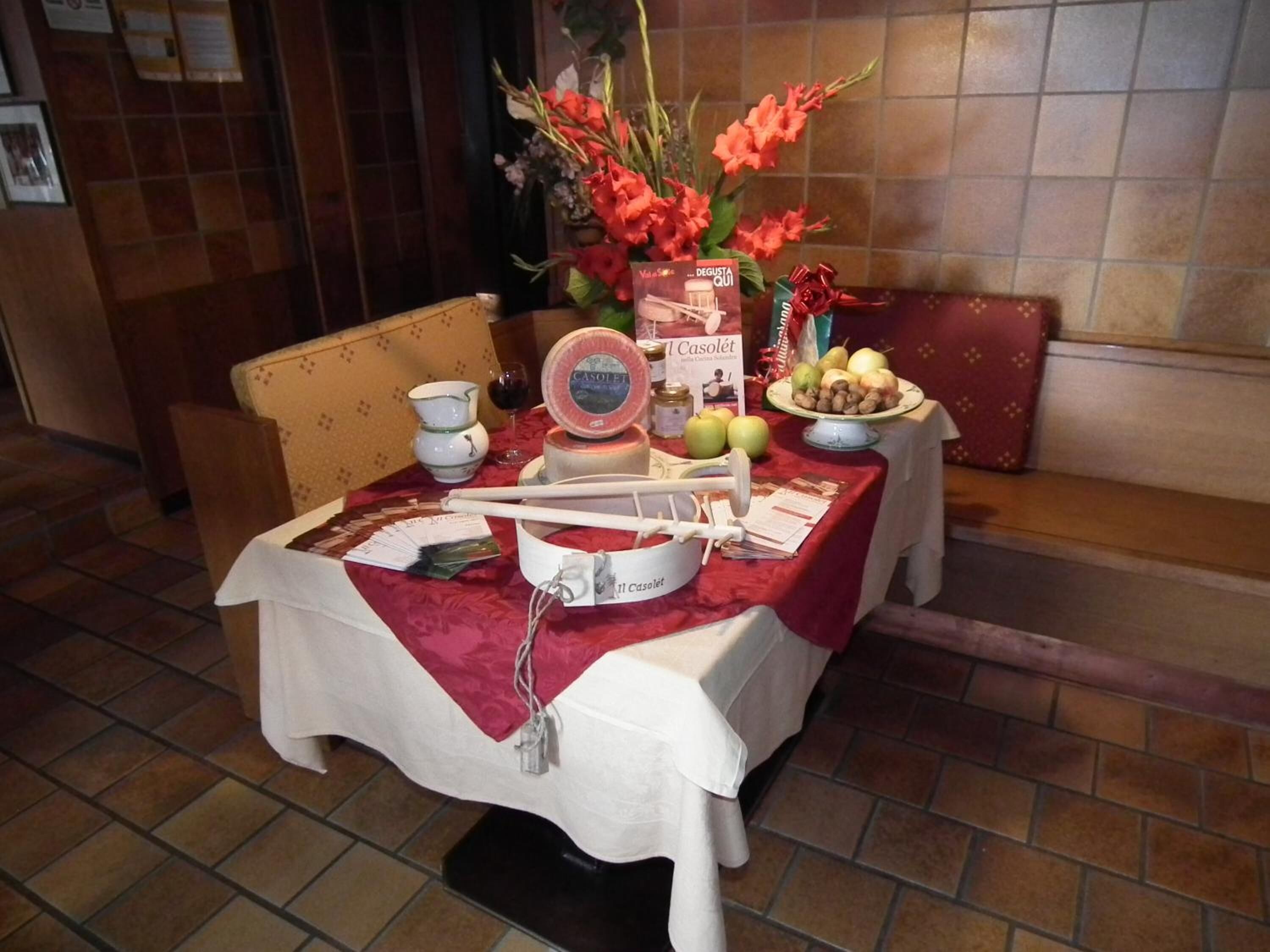 A table set with a white tablecloth, a book titled 'L'Asalet', a fruit bowl, a bottle of Casolei, and a vase with flowers.
