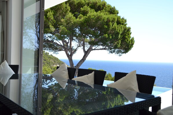 A glass table with four chairs, each with a white pillow, overlooking a tree and the ocean. - Large, Luxury Villa, Sea Views, Heated Pool in Begur, Costa Brava (La Borna)