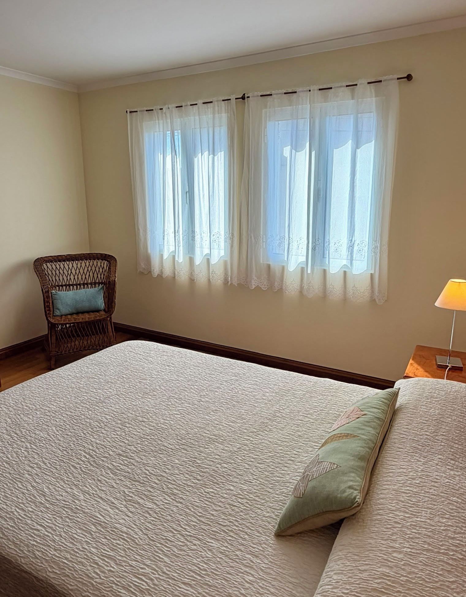 A bedroom with a large bed, a chair, a nightstand with a lamp, and a window with curtains.