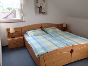 Un dormitorio con una cama de madera, dos mesitas de noche con lámparas, un ventanal con vistas a un edificio y un cuadro en la pared.
