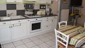 A compact kitchen with white cabinets, a gas stove, a sink, and a dining area with a striped tablecloth. - Quiet 40m² cottage with private courtyard (La Tour-Saint-Gelin)