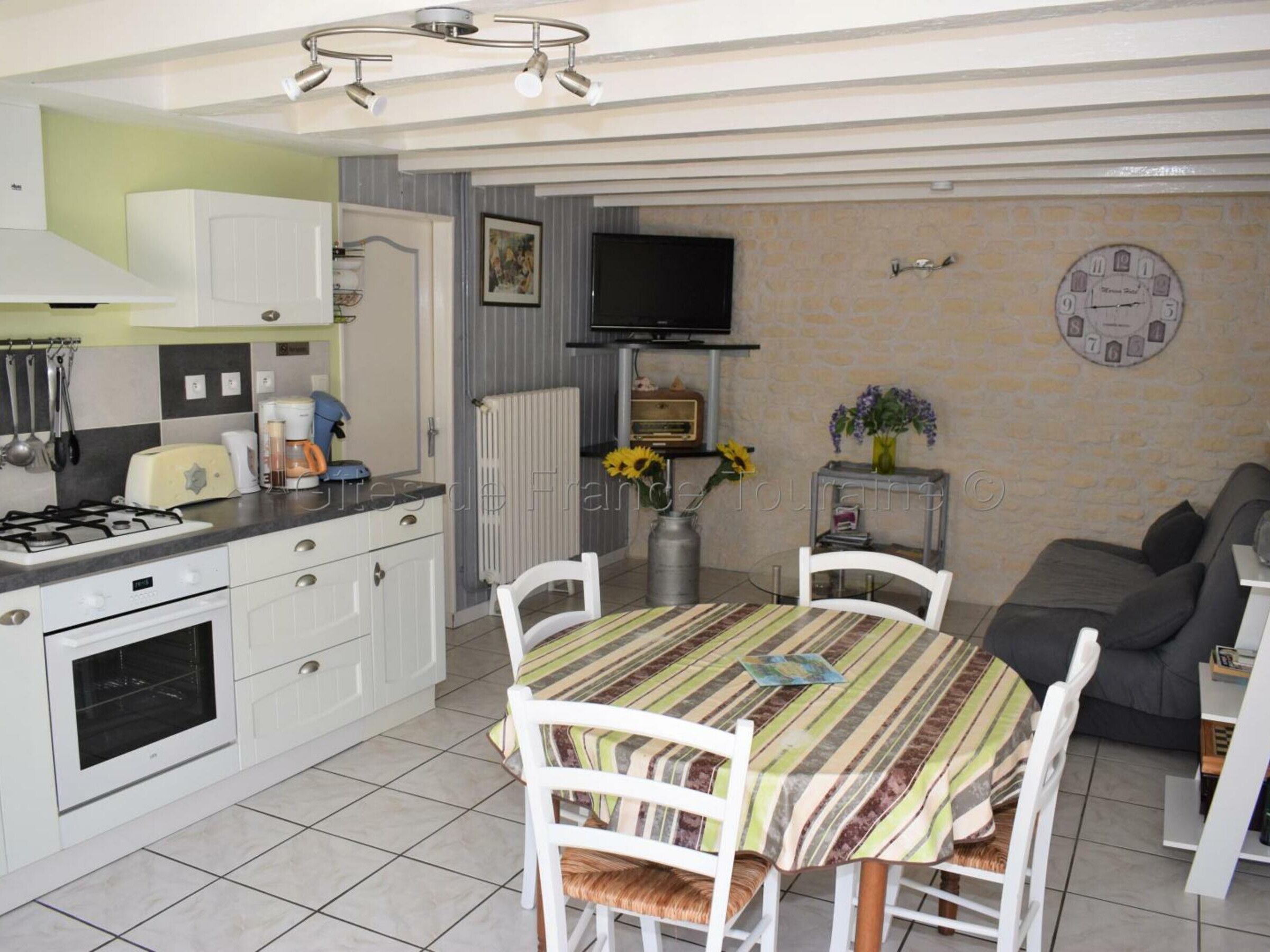 Une cuisine compacte avec des meubles blancs, une cuisinière à gaz et un four encastré. On y trouve une table à manger avec des nappes à rayures et des chaises blanches. Un téléviseur est fixé au mur, au-dessus du plan de travail de la cuisine.
