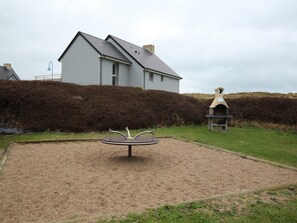 Een huis met een grijs dak en witte muren, een grindpad met een metalen sculptuur, en een haag op de achtergrond.