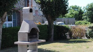 Un barbecue en pierre installé à l’extérieur, doté d’une cheminée, et situé dans un espace verdoyant agrémenté d’arbres, avec un bâtiment visible au loin.