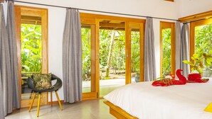 A bedroom with a large bed, a chair, and a view of greenery through open doors.