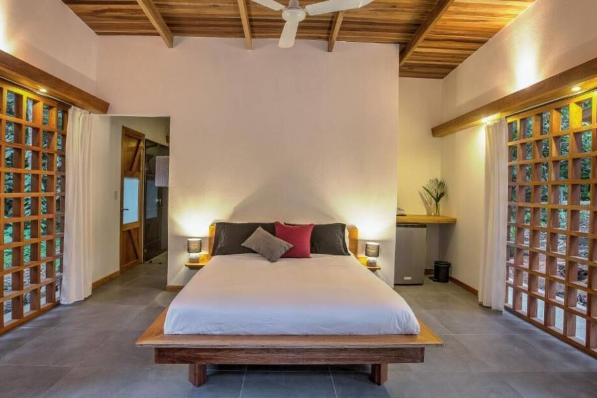 Une chambre spacieuse avec un grand lit, une tête de lit en bois et un ventilateur de plafond. On y trouve deux tables de chevet avec des lampes et une petite plante sur l’une d’elles.