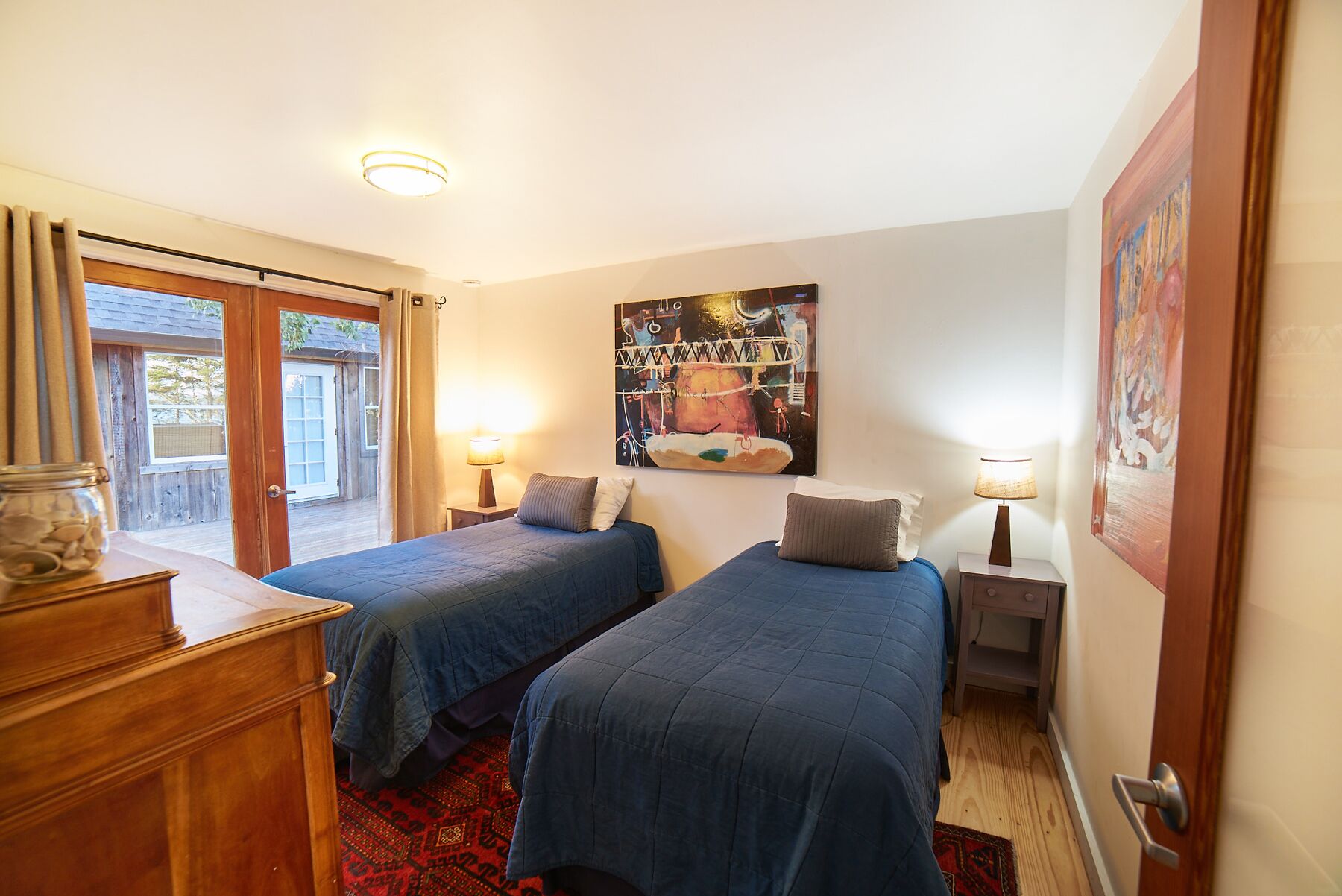 A bedroom with two beds, a wooden dresser, a painting on the wall, and a window with curtains.