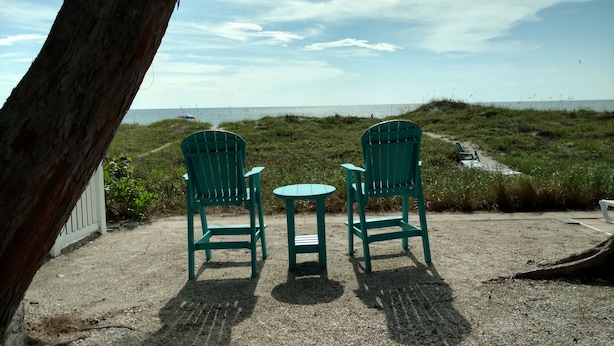 Two green chairs and a small table set on a sandy area with a view of the beach and ocean. - Beachfront Cottages PATIO at Beautiful Sandy Shores on the Gulf of America (INDIAN ROCKS BEACH)