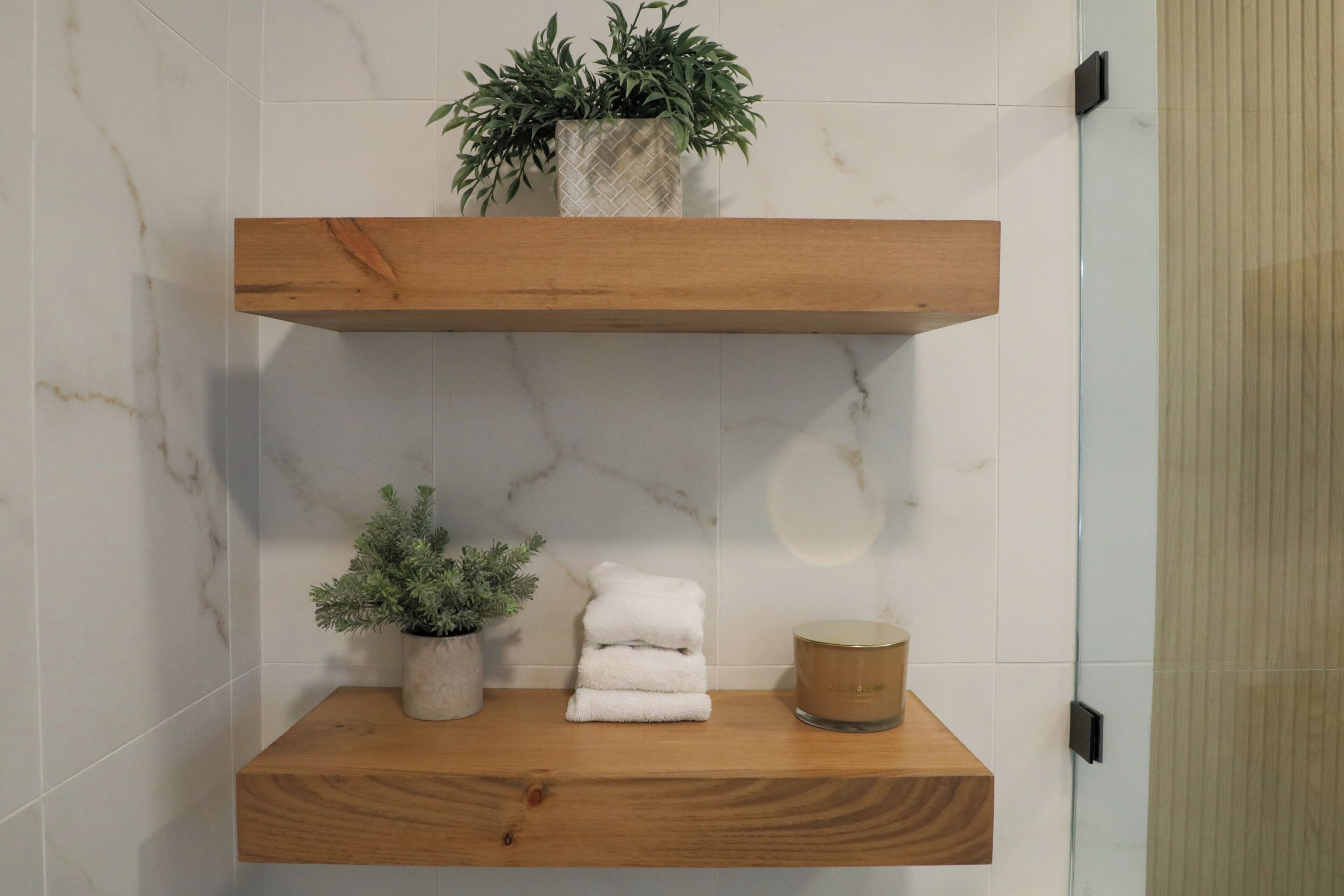 A bathroom with a wooden shelf holding a potted plant, a stack of white towels, and a candle.