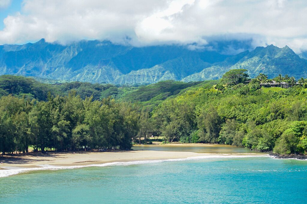 A beach with clear turquoise waters, a sandy shore, and lush green hills with a mountain range in the background.