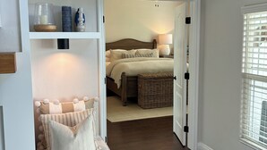 Quarto com cama, mesa de cabeceira, abajur e vista para outro cômodo por uma porta aberta.