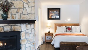 A hotel room with a stone fireplace, a bed with white bedding, a nightstand with a lamp, and a framed picture on the wall. - Schott’s Lake Conference & Resort (Sundre)