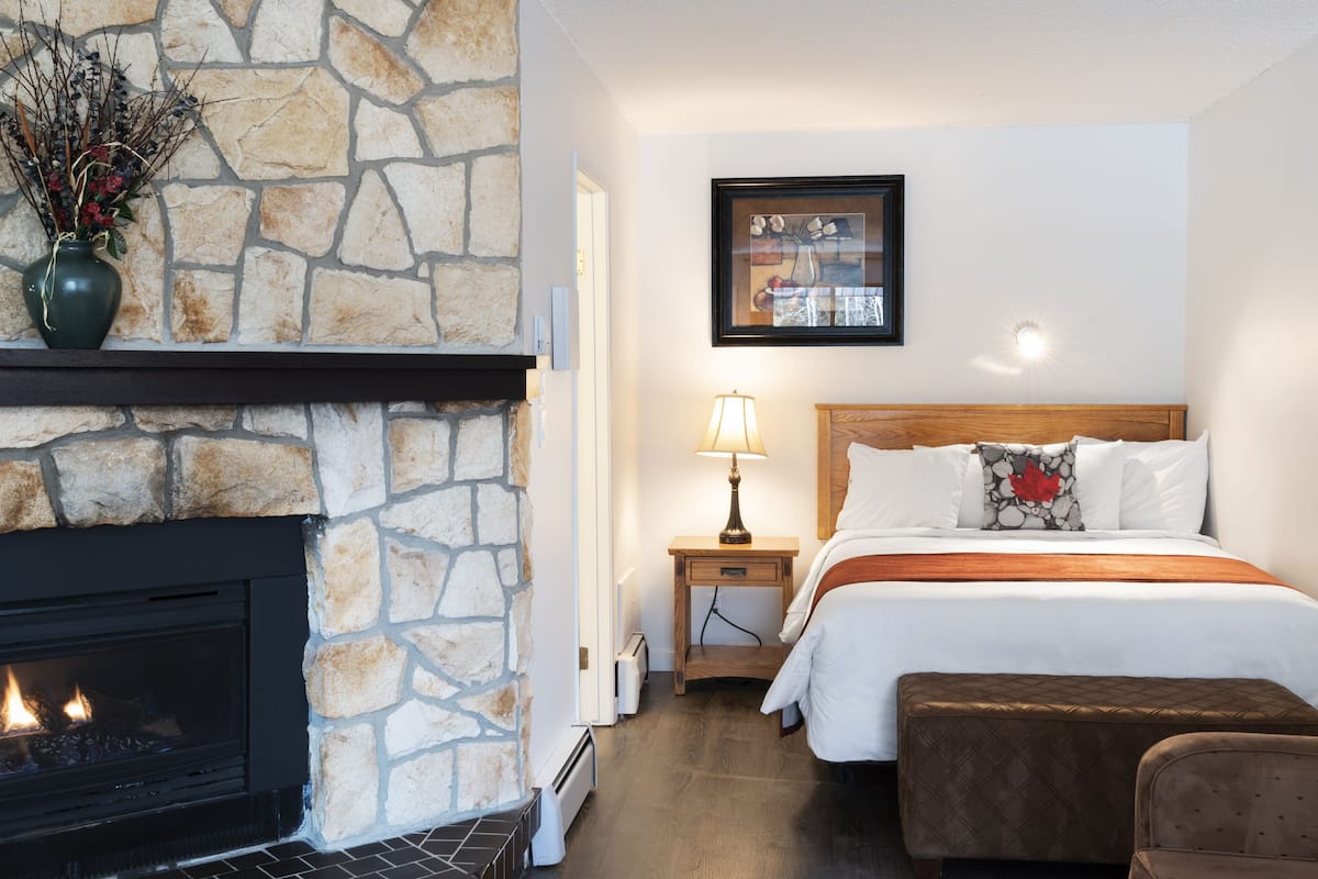 A hotel room with a stone fireplace, a bed with white bedding, a nightstand with a lamp, and a framed picture on the wall.