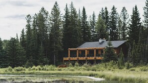 A cabin with a deck, surrounded by tall trees and a pond. - Schott’s Lake Conference & Resort (Sundre)