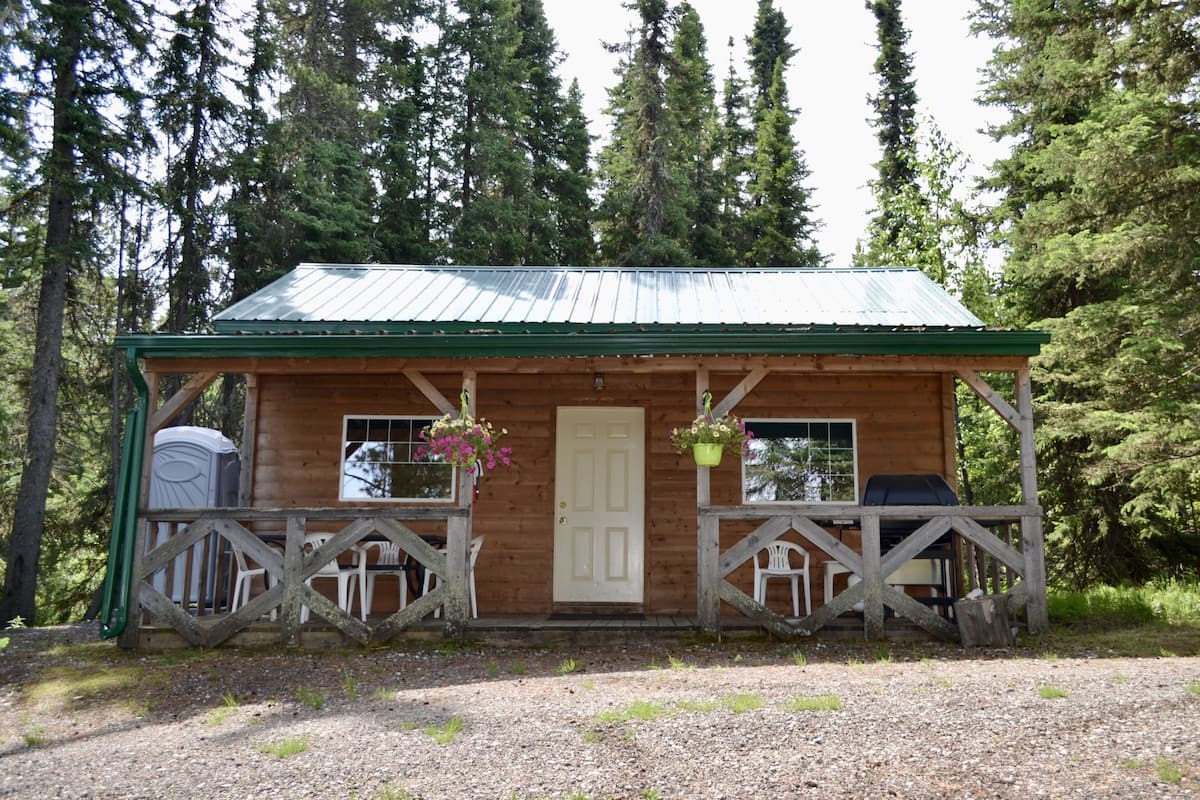 A wooden cabin with a metal roof, a porch with white chairs, and hanging flower baskets.