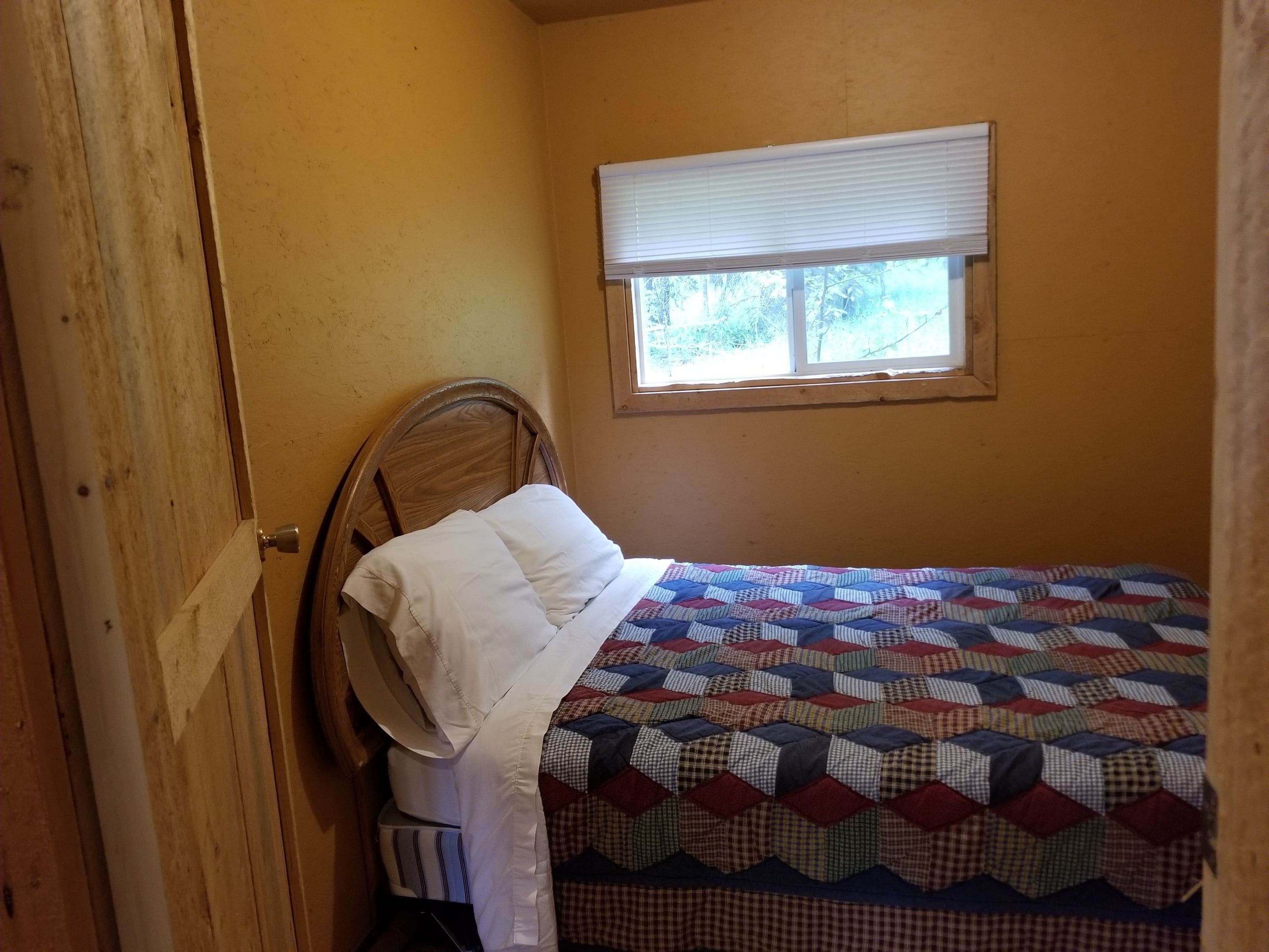 A small bedroom with a wooden headboard, a quilted bedspread, and a window with blinds.