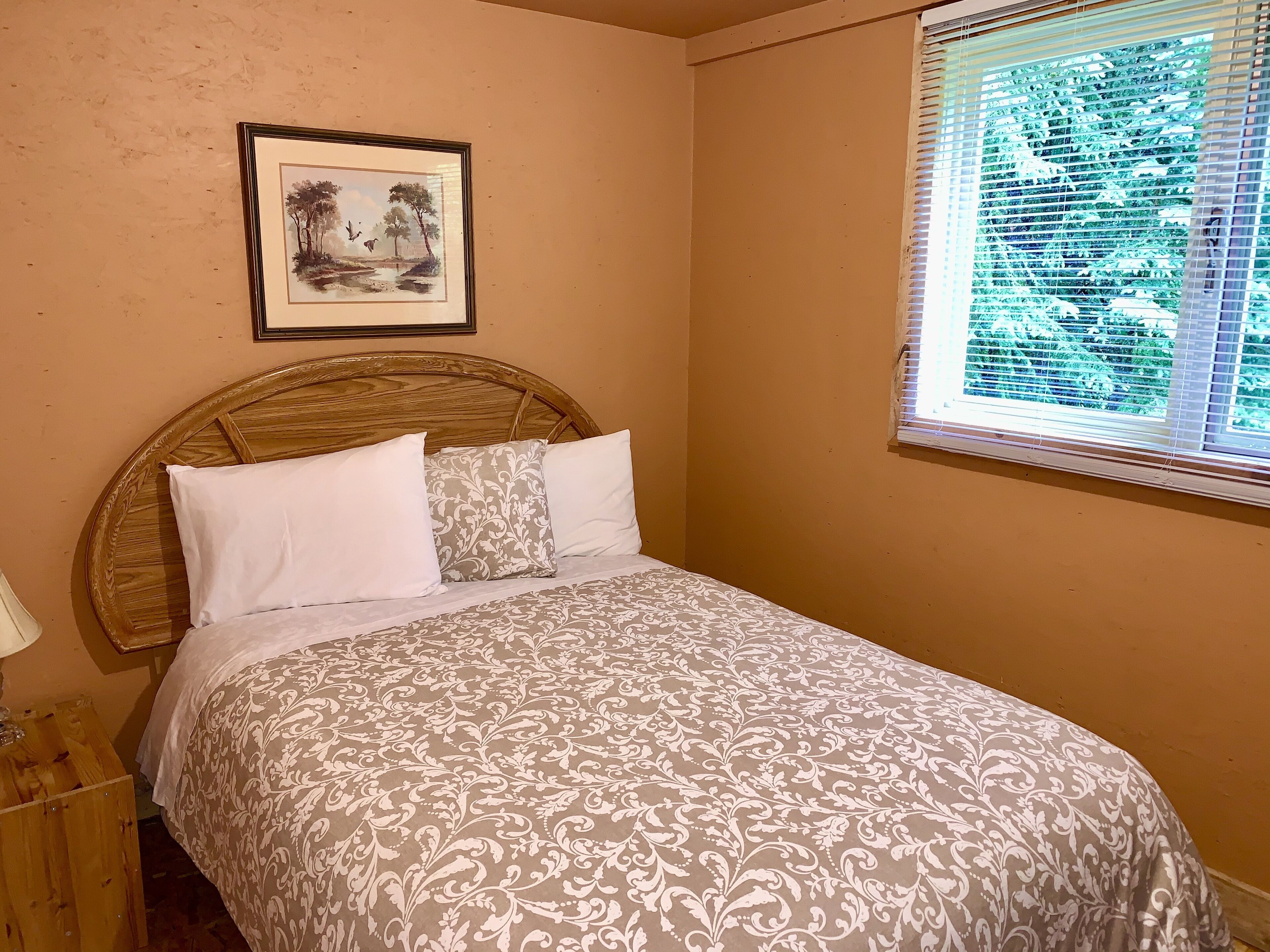 Un dormitorio con cabecera de madera, cama con ropa de cama estampada, un cuadro enmarcado en la pared y una ventana con persianas.