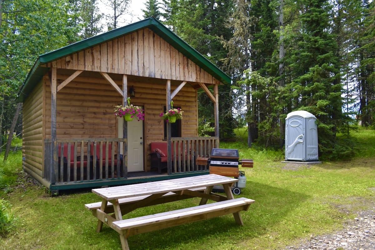 A wooden cabin with a porch, a picnic table, and a portable toilet.