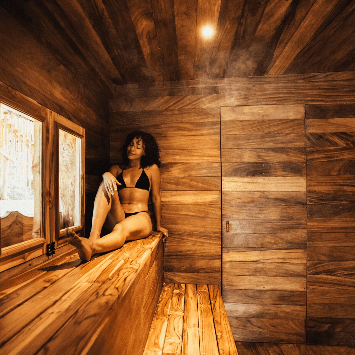 A wooden cabin interior with a person seated on a bench, a window, and wooden walls.