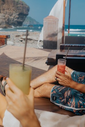 A person relaxing on a beach chair with a drink, overlooking the ocean and a rocky cliff.