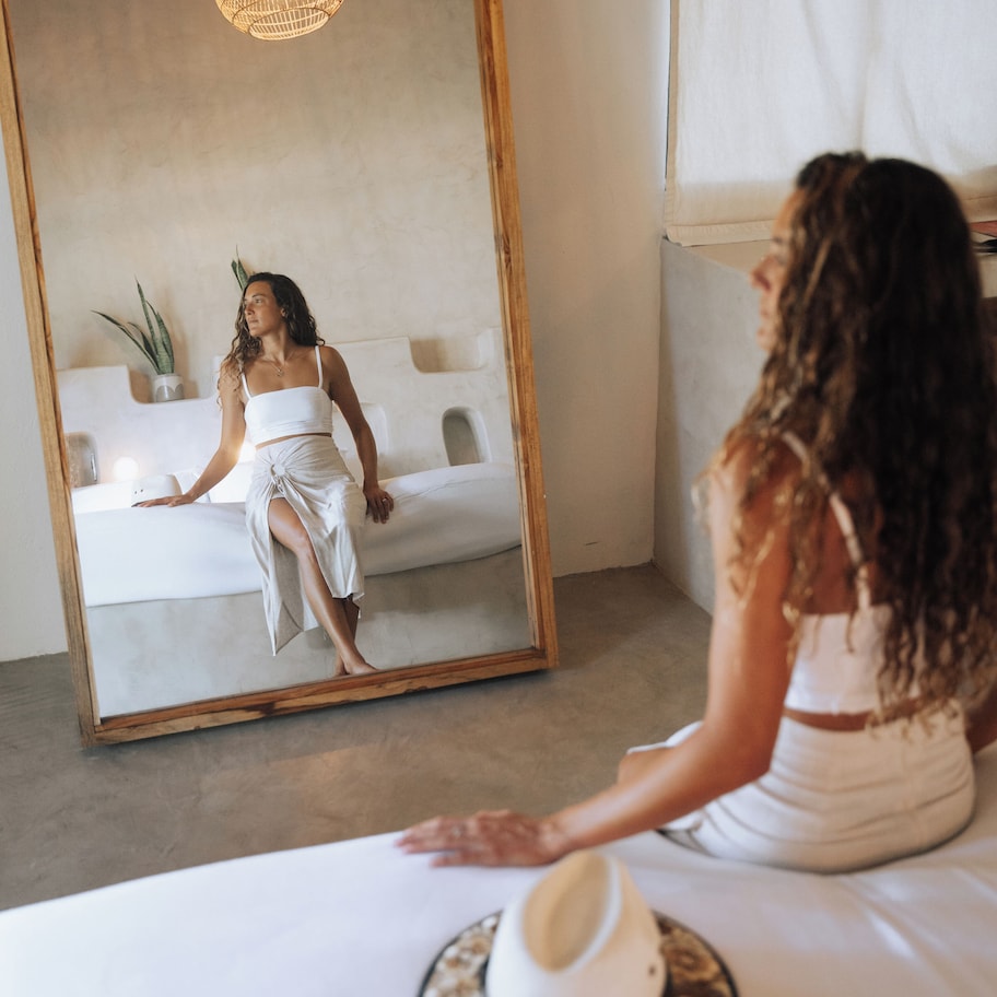A room with a large mirror reflecting a person sitting on a white couch. There is a potted plant on the wall and a wooden-framed mirror on the floor.
